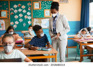 Black female teacher and schoolboy wearing face masks while using digital tablet during a class in the classroom. - Powered by Shutterstock