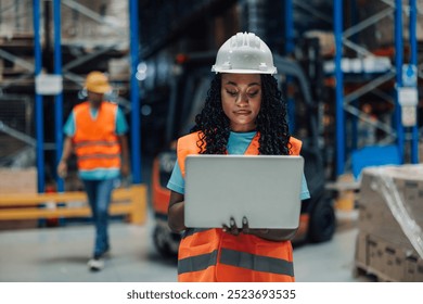 A black female warehouse worker in a hard hat and safety vest focuses intently on a laptop, showcasing technology's integration with modern logistics and safety practices. - Powered by Shutterstock