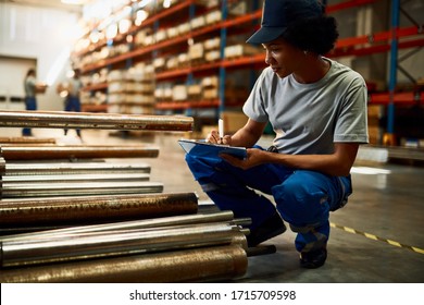 Black Female Worker Writing On Clipboard While Inspecting Steel Products At Industrial Warehouse. 