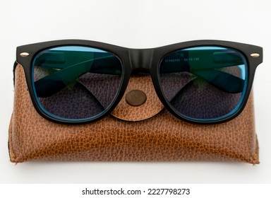 Black frame blue sun-glass made of acetate material resting on a brown leather case on white isolated background. Selective Focus in center.