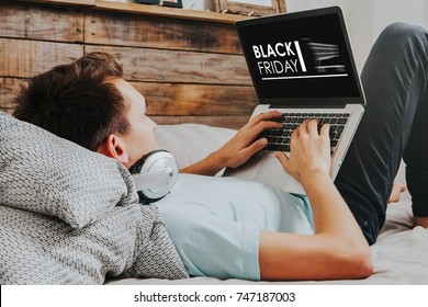 Black Friday banner in a laptop computer while man uses it to buy by internet lying down at home.