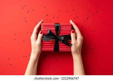 Black friday concept. First person top view photo of female hands giving red giftbox with black ribbon bow over confetti on isolated red background
