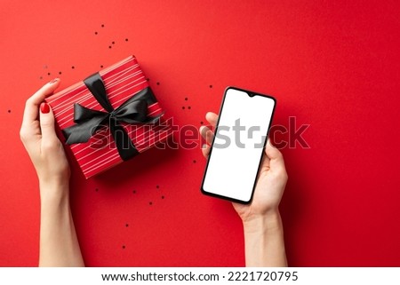 Black friday online shopping concept. First person top view photo of female hands holding smartphone and big red giftbox with ribbon bow over confetti on isolated red background with blank space