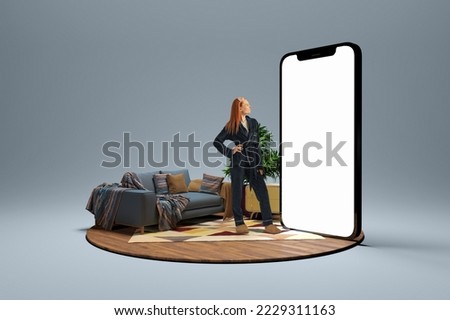 Black friday sales. Young girl in homewear standing near to 3d model of cellphone with blank white screen and dreaming isolated on grey background. Online shopping, choice, ad, new app
