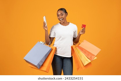 Black Friday vibes. Black woman in casual using her phone and credit card, she shops online on yellow studio backdrop, scoring discounts with style, blending fashion and tech. Shopping application - Powered by Shutterstock