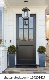 A Black Front Door Flanked By Two Potted Plants In Blue Pots. The House Is White. Also Seen Is A Light Fixture, Off, And Part Of A Large Window. A Stone Porch Leads To The Door And Doormat.