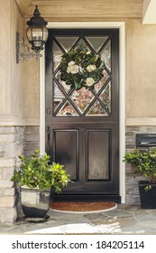 Black Front Door To Home With Flower Wreath. A Black Front Door To A Home With An Ornate Flower Wreath And Classic Light Fixture. Also Seen Are Door Plants, And A Stone Porch. 