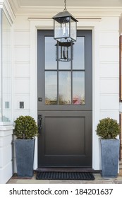 A Black Front Door To A White Family Home In Daytime With Door Mat, Light Fixture, Plants In Blue Planters, And A Hint Of A Large Window.