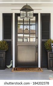 A Black Front Door Of A White Family Home With Gray Porch. The Door Is Framed By Black Planters, Black Decorative Shutters, And A Light Fixture. Also Seen Is A Duck Figurine, And A Porch Chair.