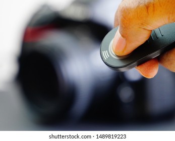 A Black Full Frame Digital SLR Camera In The Background, And A Man's Fingers Holding And Pressing The Switch Of Wireless Infrared Remote Control Device In The Foreground.                           