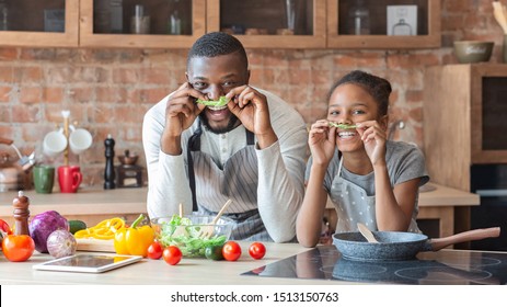 Black funny father and daughter making mustaches with lettuce, having fun while cooking at kitchen, panorama with empty space