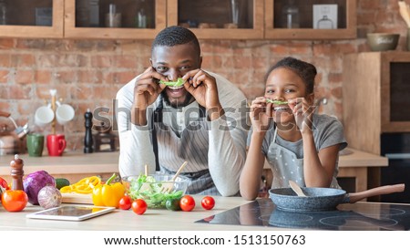 Black funny father and daughter making mustaches with lettuce, having fun while cooking at kitchen, panorama with empty space