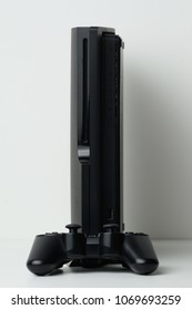 Black Game Console Is Stand On The White Table With Game Pad.