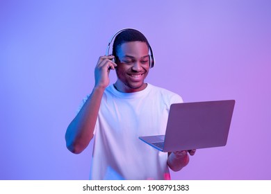 Black Gamer Man In Wireless Headset Playing Video Games On Laptop, Looking At Computer Screen While Standing In Vivid Neon Light Over Purple Studio Background, Guy Enjoying Cyber Gaming, Copy Space