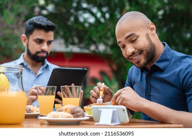 Black Gay Couple Having Breakfast And Looking At Mobile Outside In The Garden