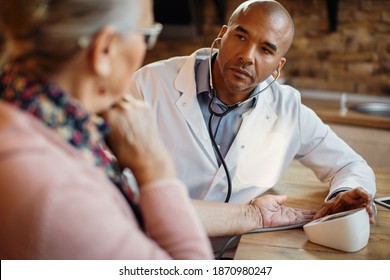 Black general practitioner measuring blood pressure of mature woman during home visit. 