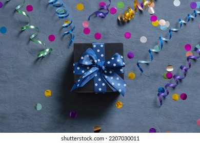 Black Gift Box With Bow And Holiday Tinsel Flat Lay On Dark Cement Background. Top View.