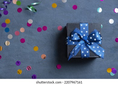 Black Gift Box With Bow And Holiday Tinsel Flat Lay On Dark Cement Background. Top View.