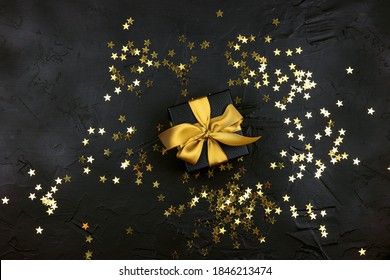 Black Gift Box With Gold Ribbon And Gold Star Sequins On A Black Background. Holiday Or Black Friday Concept. Top View With Copy Space.