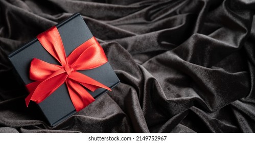 Black Gift Box With Red Satin Ribbon On Dark Velvet Background With Copy Space. Top View, Minimalistic Present