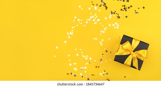 Black gift box with yellow bow on yellow background with sparkling confetti. Flat lay.