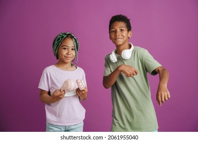 Black girl and boy with headphones smiling and dancing at camera isolated over purple background