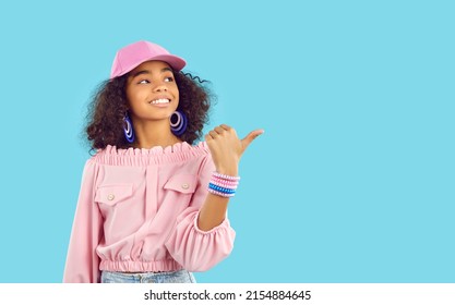 Black Girl With Curly Hair Wearing Pink Baseball Hat, Blouse, Bracelet And Earrings Standing And Pointing Sideways At Copy Space On Blue Background. Modern Kids Fashion, Child Model Studio Concept