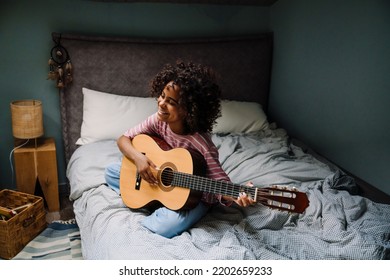 Black Girl Laughing And Playing Guitar While Sitting On Bed At Home