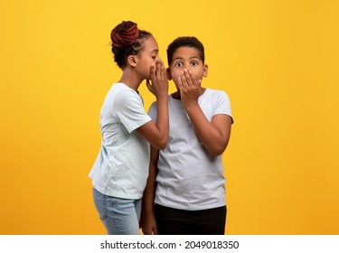 Black Girl Sister Sharing Secrets With Her Brother, African American Teen Girl Whispering Something To Cute Boy Sibling, Yellow Studio Background. Brotherhood, Sisterhood, Friendly Family Concept