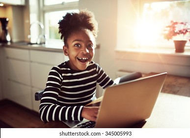 Black girl sitting playing on a laptop computer at home looking at the camera with a joyful expression of amazement and wonder