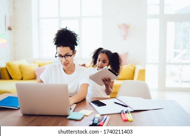 Black Girl Standing In Bright Room And Showing Tablet To Mother While Parent In Glasses Working In Front Of Laptop And Concentrating