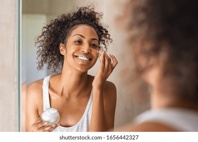 Black girl take care of her beautiful skin. Young african woman applying moisturizer on her face. Smiling black natural girl holding little jar of skin lotion in bathroom for beauty treatment routine. - Powered by Shutterstock