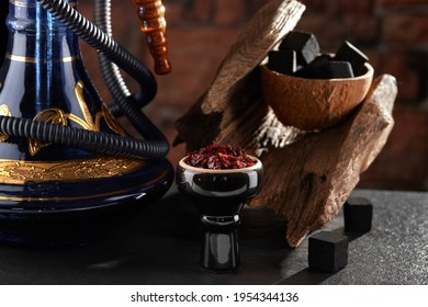 Black glossy hookah bowl filled with apple shisha and prepared for smoking stands on a dark surface next to a beautiful hookah, charcoal and textured wood. High quality photo.