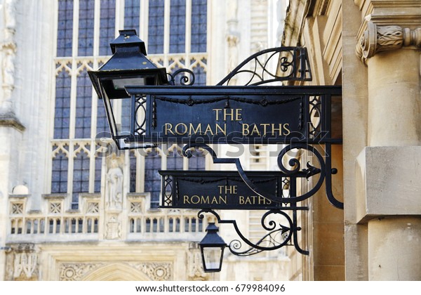 Black and gold signs on decorative\
metalwork brackets for The Roman Baths at Bath Spa, Somerset, UK\
showing Bath Abbey in the background and a traditional\
lamp