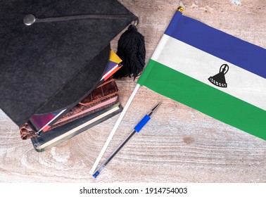 Black Graduation Hat On Books Next To Lesotho Flag, Education Concept, Top View