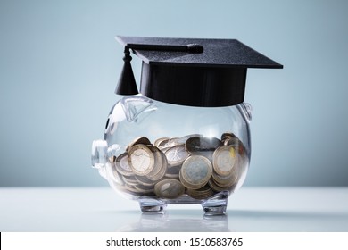 Black Graduation Hat With Transparent Piggy Bank Arranged On White Desk Over Blue Backdrop