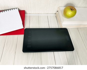 Black graphics tablet with notebook and stack books on white wooden desk. Digital art, design, illustration, creative work, drawing, education, study workspace setup with copyspace