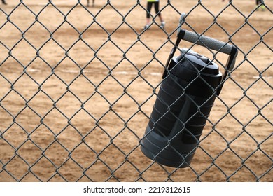 Black And Gray Large Water Jug Hangs On A Chain Link Fence. Stay Hydrated During Team Sports.