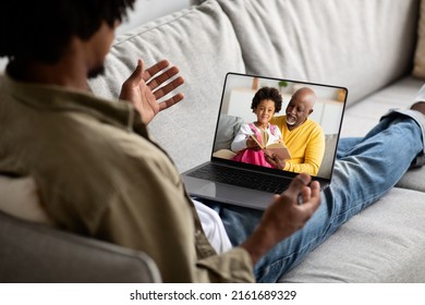 Black Guy Calling His Family Senior Man And Baby Child While Staying Apart, Reclining On Sofa At Home And Using Laptop, Looking At Computer Screen And Gesturing, Over Shoulder Shot