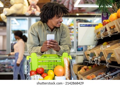 Black Guy Doing Grocery Shopping Using Cellphone Buying Food Standing With Shop Cart In Supermarket On Weekend. Technology And Commerce, Mobile Offer Concept