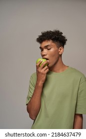 Black Guy Eating Fresh Green Tasty Organic Apple. Concept Of Dieting And Healthy Eating. Young Brunette Curly Man Wearing T-shirt. Isolated On Grey Background. Studio Shoot. Copy Space