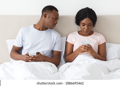 Black Guy Eavesdropping On His Girlfriend Using Mobile Phone, Looking At Her Smartphone Screen. African American Young Couple Sitting In Bed And Using Mobile Phones. Distrust In Relationships