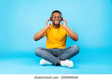 Black Guy Listening To Music With Eyes Closed Wearing Wireless Headphones Sitting In Lotus Position Over Blue Studio Background. Musical Application For Online Meditation. Relaxation Concept