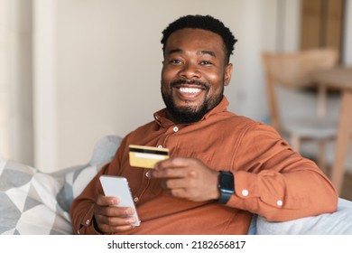 Black Guy Shopping Online Using Mobile Phone And Credit Card Sitting At Home, Smiling To Camera. Happy Man Making Payment Via Financial Application. Internet Banking App Concept