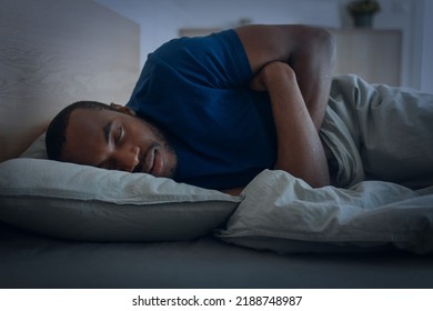 Black Guy Sleeping Lying Under Blanket In Bedroom At Home At Night. Male Resting Napping Peacefully In Bed. Recreation And Relaxation Concept. Low Light