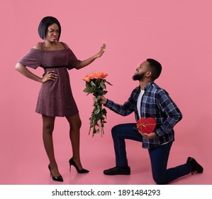 Black Guy Standing On One Knee With Flowers And Gift For Valentine's Day, Young Woman Rejecting Him On Pink Background. Offended Lady Refusing Her Boyfriend's Present After Fight, Full Length Portrait