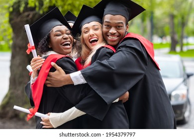 Black guy and two young ladies in graduation costumes posing at camera at university campus, holding diplomas, laughing and hugging, having happy graduation day, closeup portrait