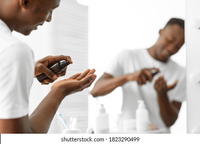 Black Guy Using Aftershave Lotion Caring For Facial Skin After Shaving Standing Near Mirror In Bathroom At Home. Male Skincare Cosmetic Products, Self-Care Routine For Men. Selective Focus