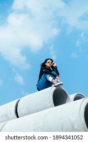Black hair girl in sweater and jeans sitting on the top of big concrete tube with knees up and grab her sunglasses.