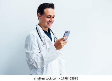 Black Haired Doctor Wearing White Coat And Stethoscope Looking At His Mobile Phone And Smiling While Making A Video Call, On White Background. Medicine Concept